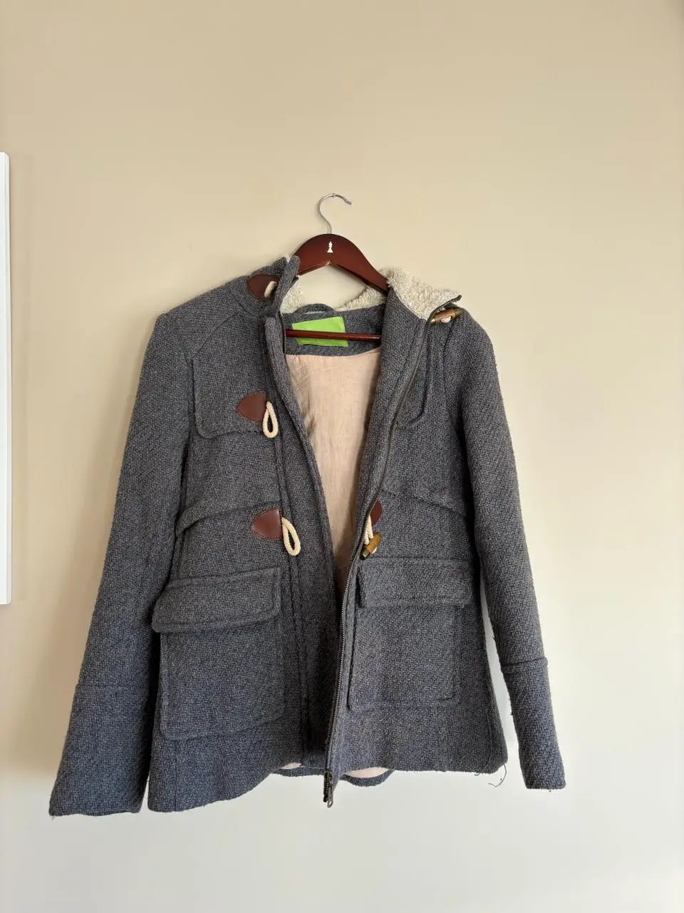 Campera de abrigo tipo duffle coat, gris con detalles en marrón. Posee capucha con forro de piel sintética, bolsillos amplios y cierre con botones de madera y tientos. Ideal para el invierno.
