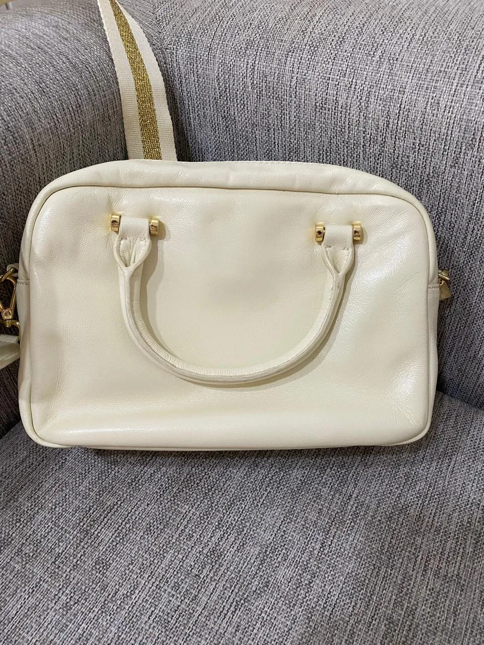 Bolso blanco de cuero con detalles acolchados y correa dorada con detalles de rayas blancas. Tiene un compartimento principal con cierre y un bolsillo interior.