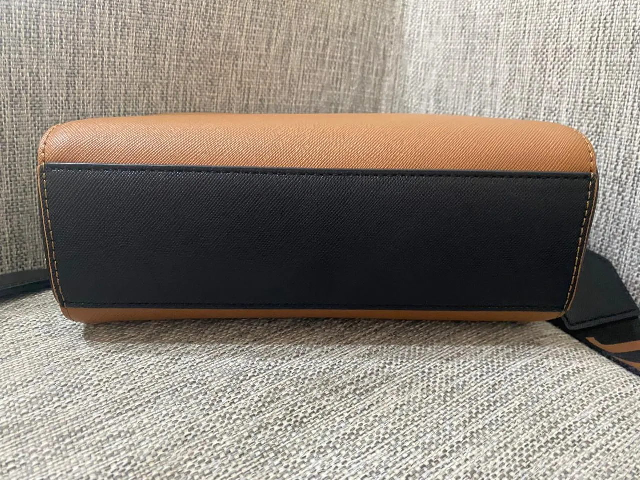 Bolso de cuero color suela con detalle de cuero negro en la base. Correa de tela negra con logo de la marca en color suela.