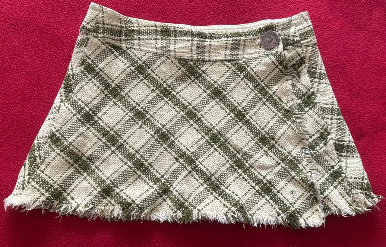 Pollera mini de tela escocesa en tonos verde y blanco, con detalle de flecos en el borde inferior. Diseño cruzado con un botón decorativo.