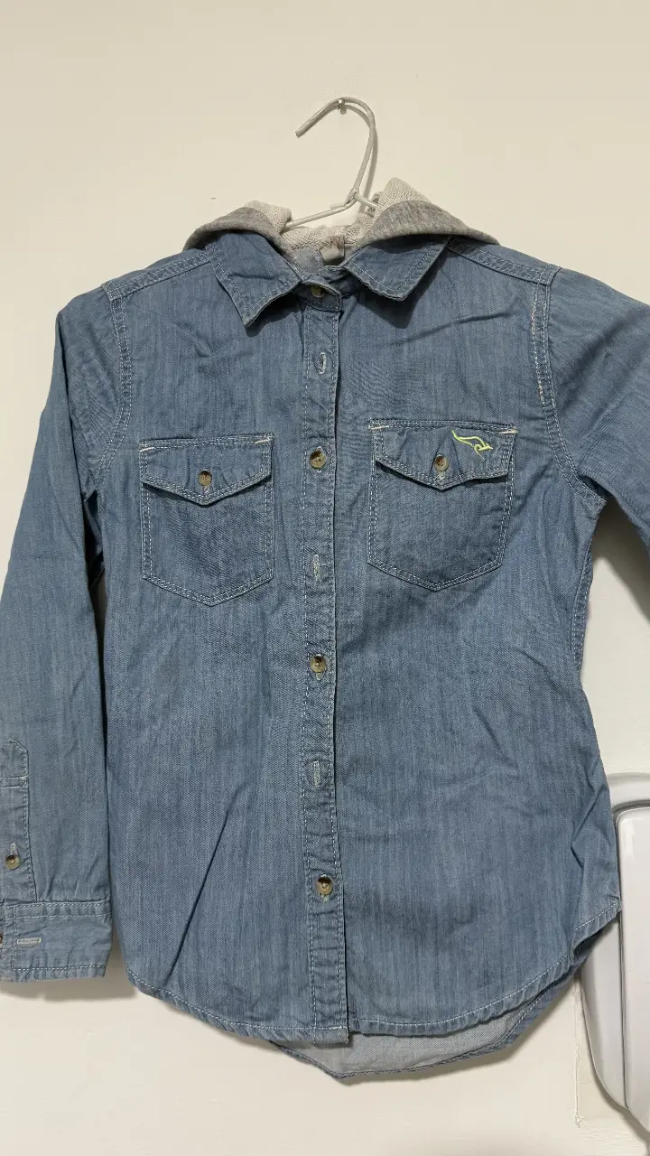Camisa de jean azul con capucha gris. Posee dos bolsillos delanteros y cierre con botones. Ideal para un look casual y canchero.