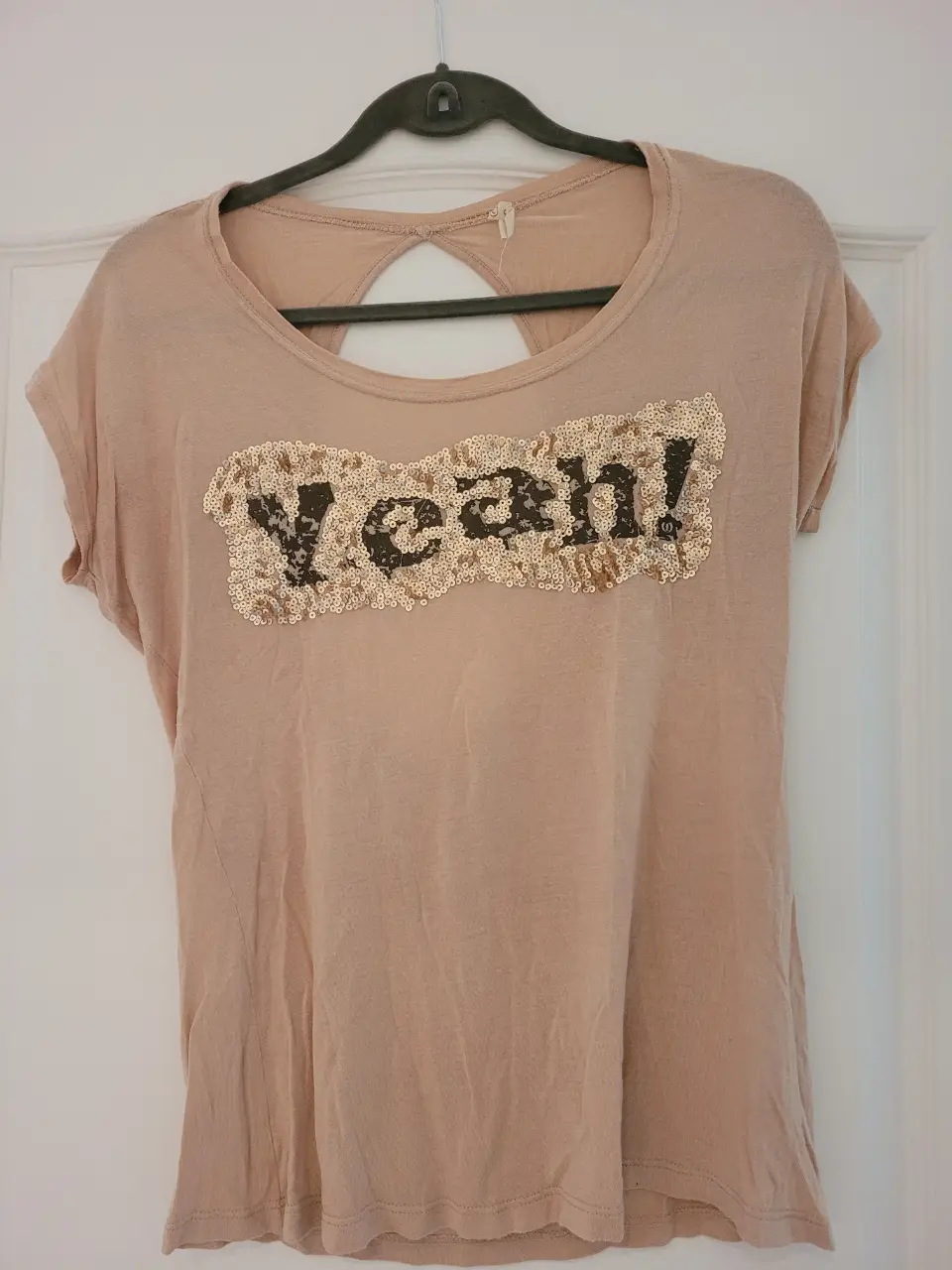 Remera de mujer color crudo con detalle de lentejuelas en el frente formando la palabra "Yeah!". Tiene un corte en la espalda.