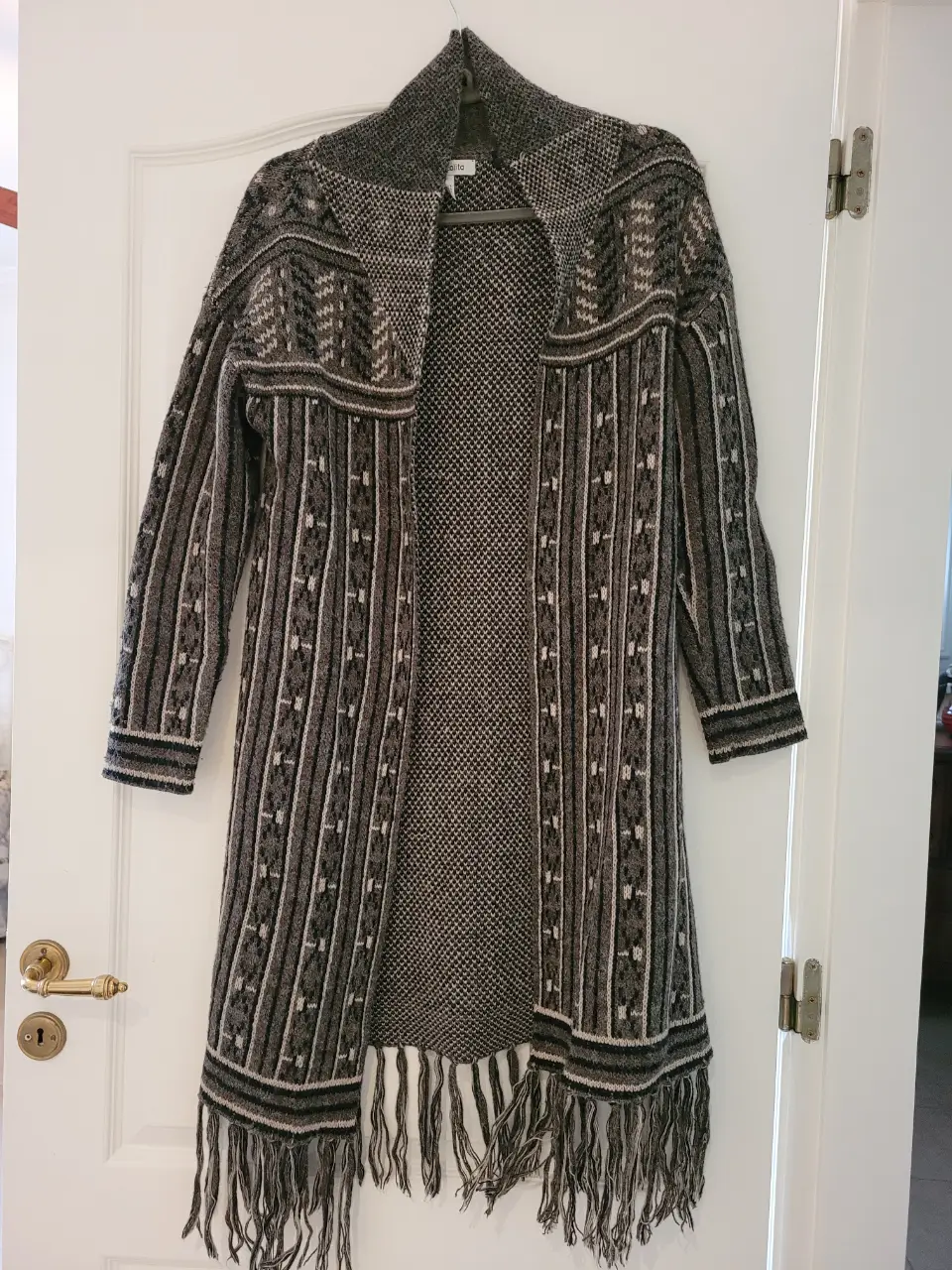Cardigan largo de tejido grueso con diseño geométrico en tonos grises y blancos, con flecos en el borde inferior. Ideal para un look bohemio y abrigado. Dice talle S pero es más M