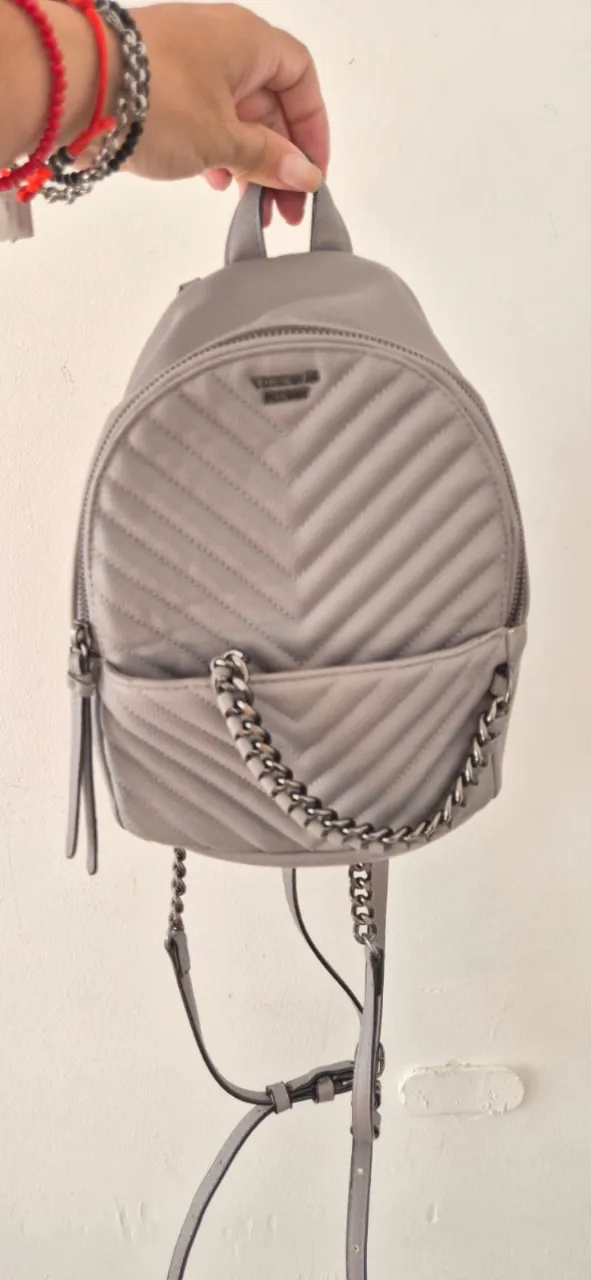 Mochila súper canchera de cuero sintético gris, con detalles de matelassé en forma de V y una cadena metálica que le da un toque rockero. Tiene un bolsillo frontal y cierre principal. Ideal para darle un toque chic a cualquier look.