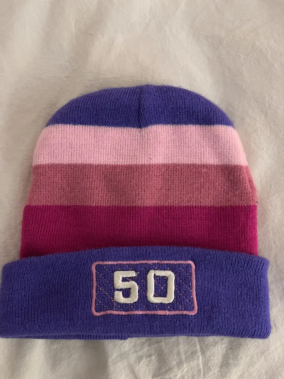 Gorro de lana con diseño a rayas en tonos violeta, rosa y fucsia. Cuenta con el número 50 bordado en la parte delantera. Ideal para un look casual y abrigado.