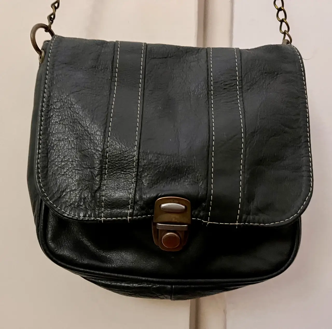 Bandolera de cuero negro con costuras blancas. Diseño clásico con solapa y cierre metálico. Ideal para un look casual y versátil. Cuero de vaca y cadena metálica.