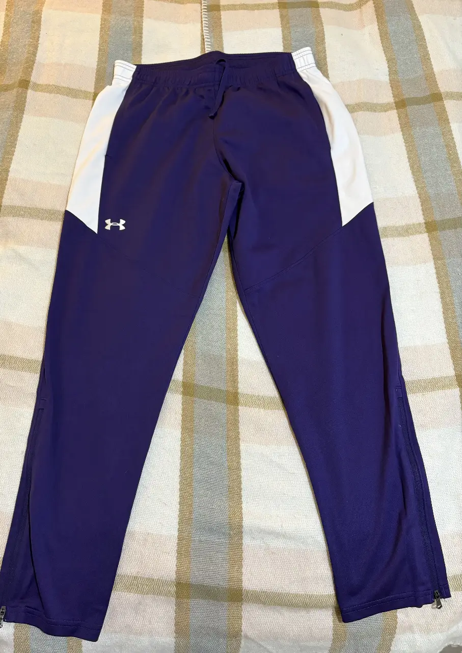 Pantalón deportivo Under Armour para hombre, color violeta con detalles en blanco. Ideal para entrenamientos y actividades físicas.
