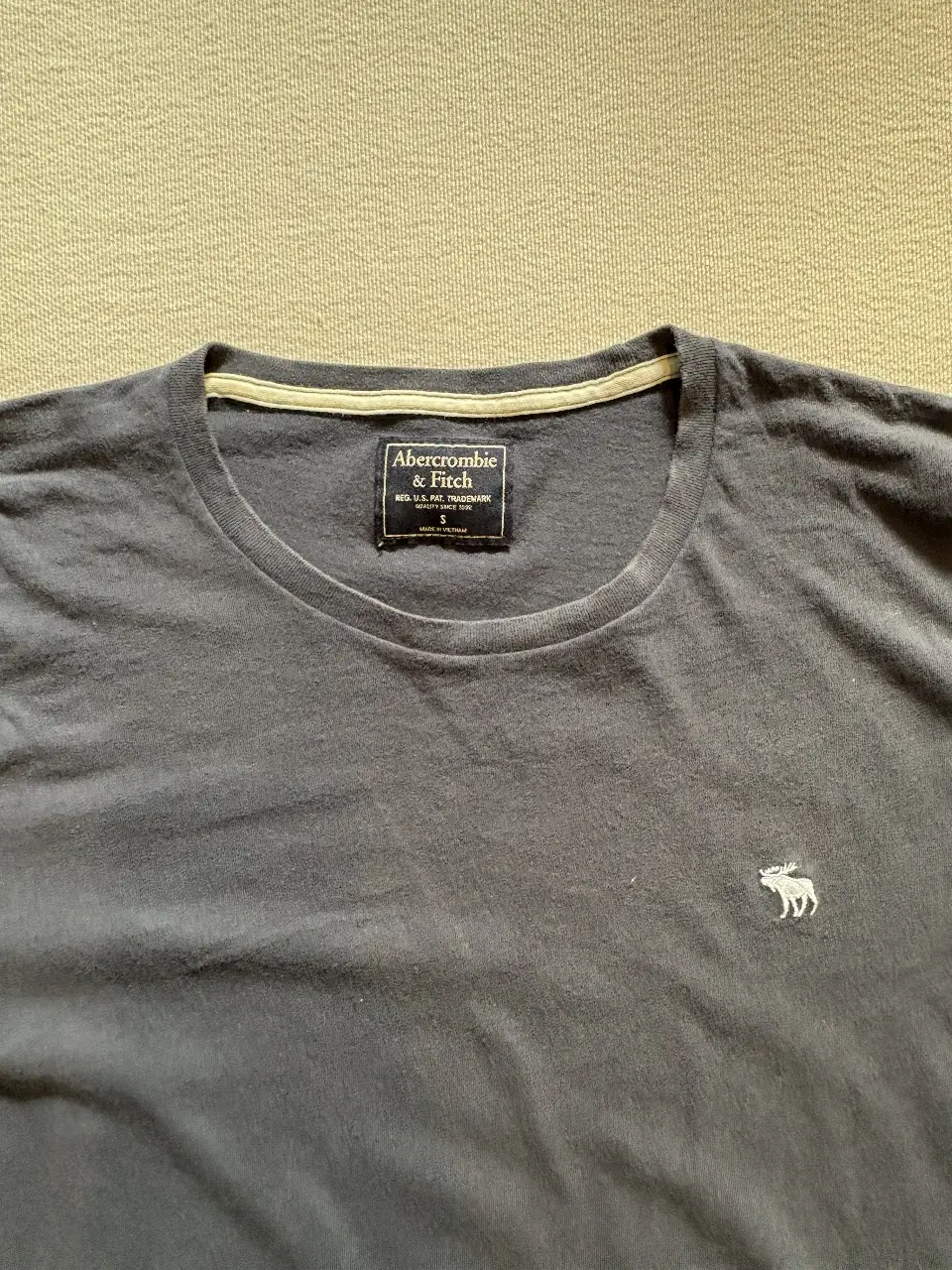 Remera de hombre Abercrombie & Fitch, de color gris oscuro, con cuello redondo y logo bordado en el pecho. Ideal para un look casual y cómodo.