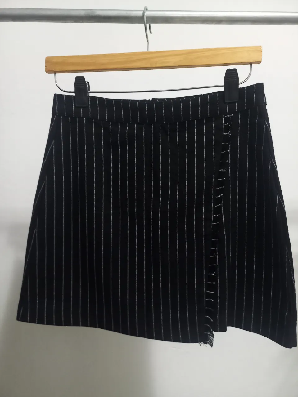 Pollera sastrera negra con rayas blancas verticales. Tiene un detalle de volado en el lateral y cierre. Ideal para un look elegante y moderno.