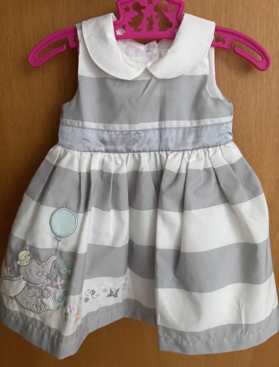 Vestido a rayas grises y blancas con un dibujo de elefante y globos en la parte inferior. Incluye un cinturón plateado y un lazo en la espalda. Viene con un cardigan plateado brillante a juego.