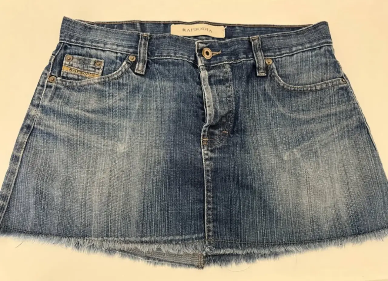 Minifalda de jean azul, con detalles de desgaste y flecos en el borde inferior. Diseño clásico de cinco bolsillos y cierre con botón y cremallera. Ideal para un look casual y moderno.