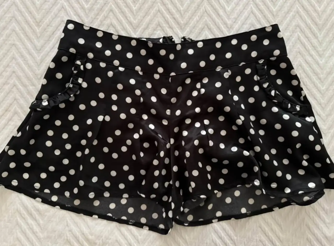 Shorts de mujer con estampado de lunares blancos sobre fondo negro. Diseño de tiro alto con detalles de volados en los bolsillos y cierre trasero. Ideal para un look de fiesta o salida.