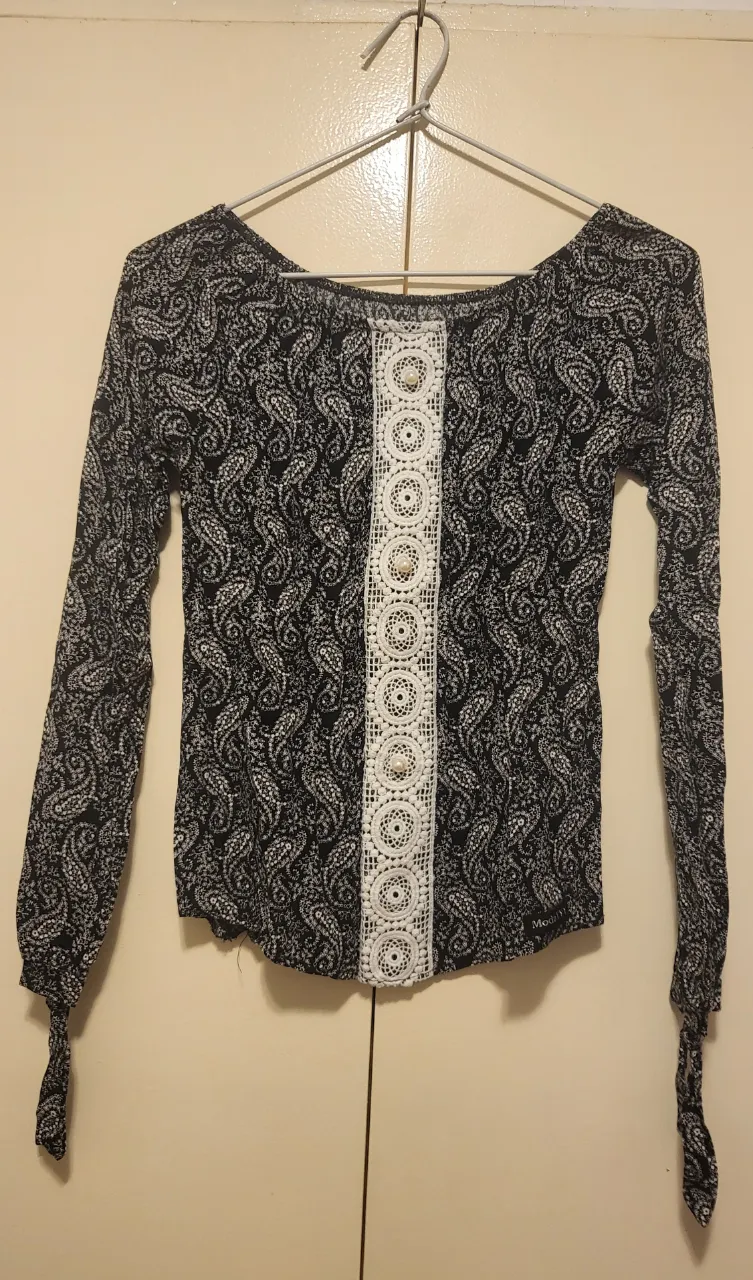 Remera manga larga estampada con diseño paisley en blanco y negro. Posee un detalle de encaje blanco con perlitas en el frente y lazos en los puños. Ideal para un look casual y canchero.