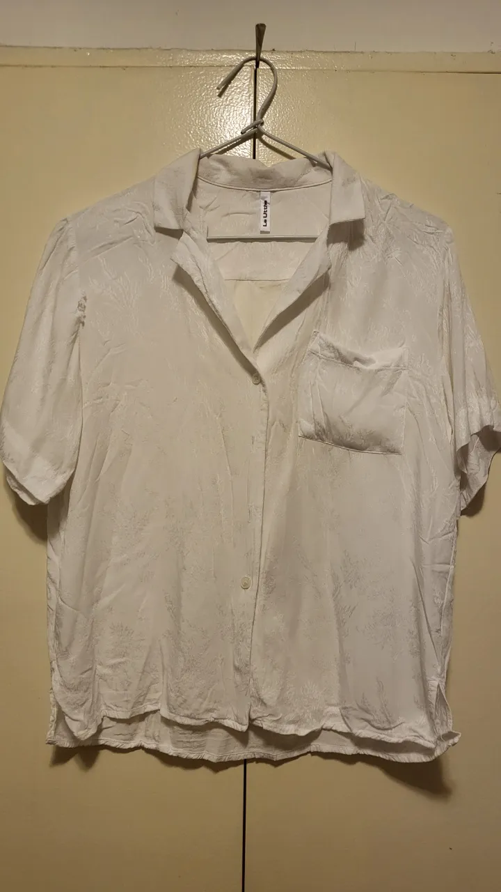 Remera blanca de manga corta con un delicado estampado floral en relieve. Posee cuello camisero, un bolsillo en el pecho y botones al frente. Perfecta para un look casual y fresco.