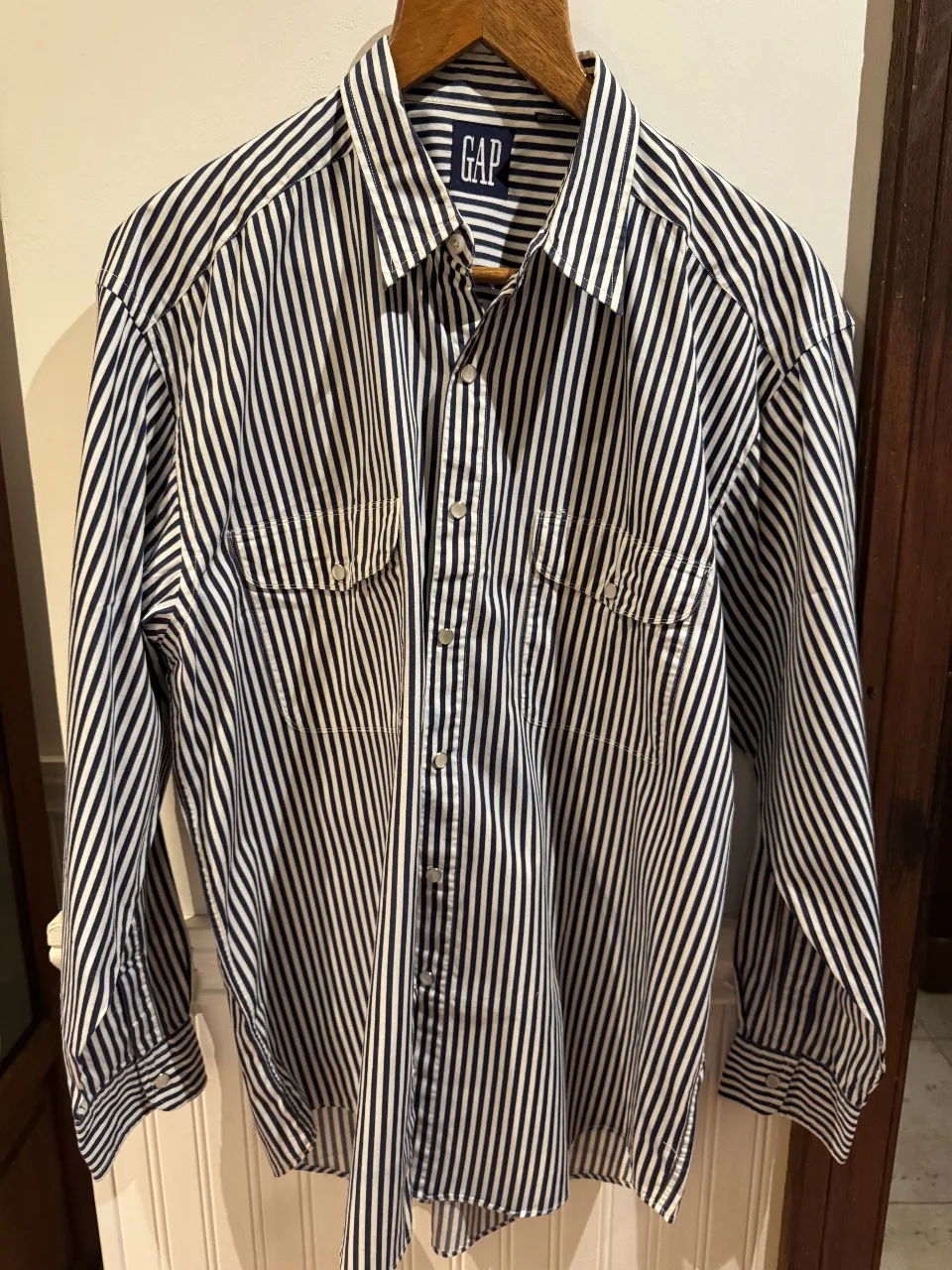 Camisa a rayas azul y blanca de hombre, de la marca GAP, manga larga, con dos bolsillos delanteros y cuello clásico. Ideal para un look casual y cómodo.