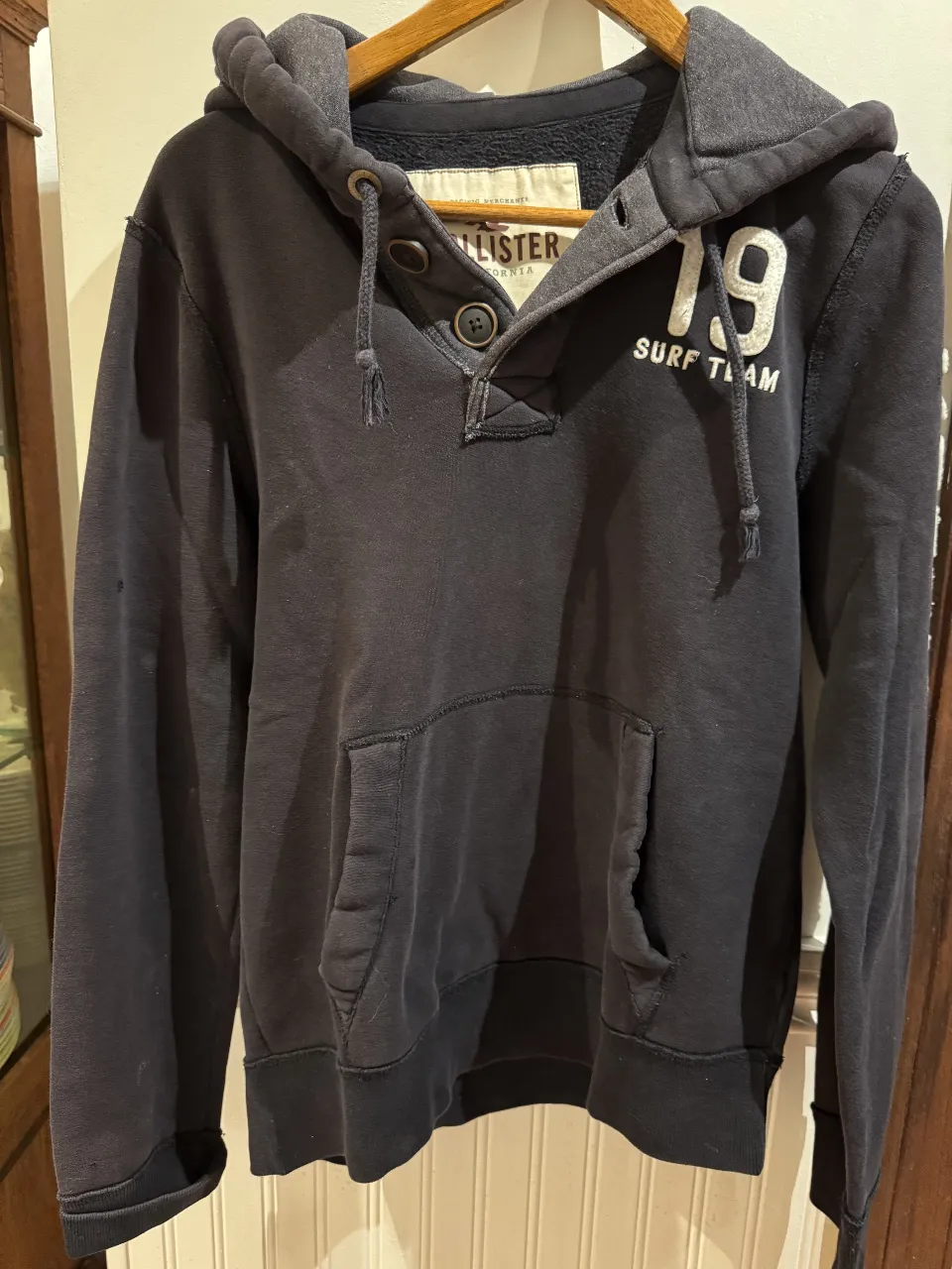 Buzo canguro Hollister negro con capucha y bolsillo delantero. Detalles en blanco y botones en el cuello. Ideal para un look casual y abrigado.