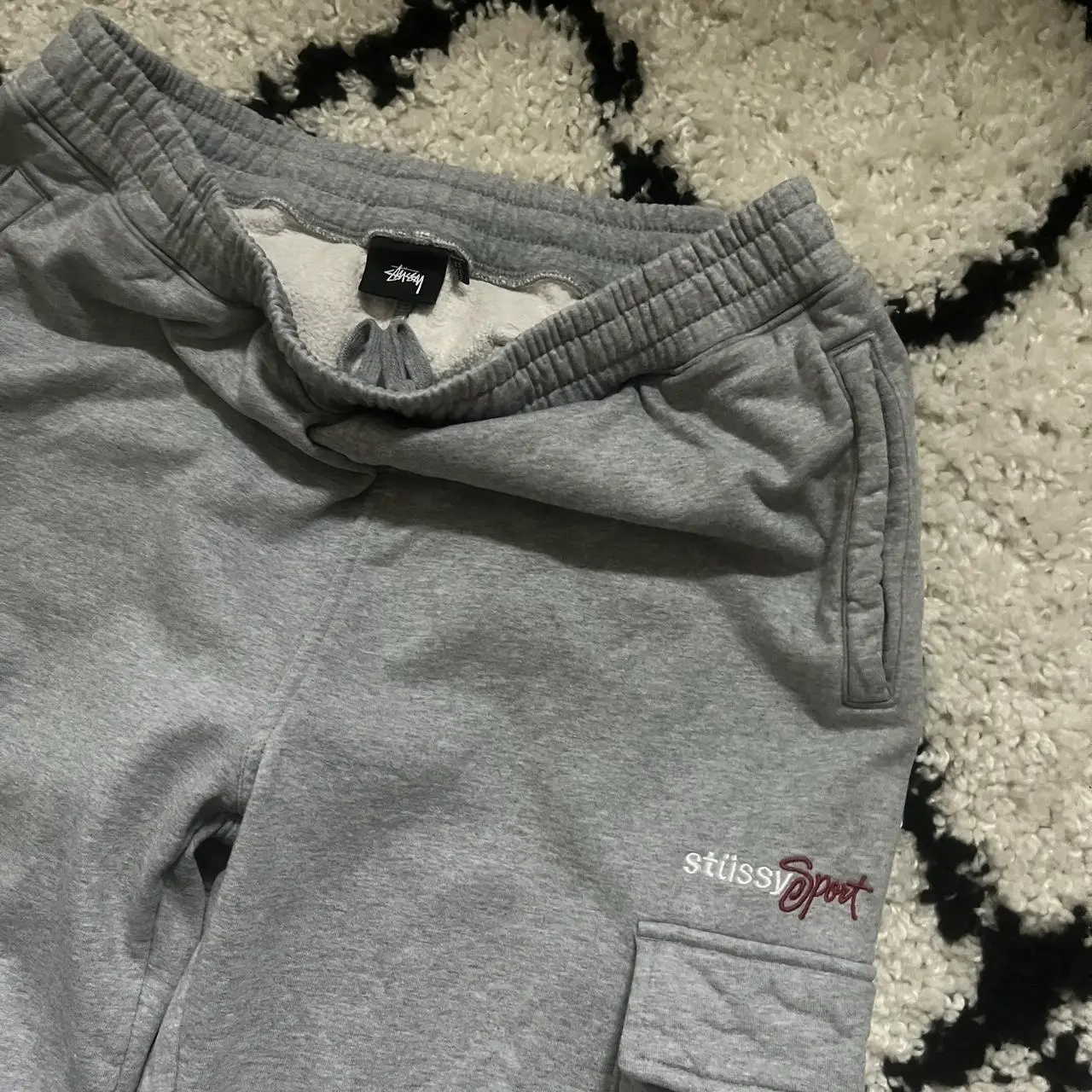 Pantalón cargo gris de hombre, con el logo "stussy sport" bordado en rojo. Posee bolsillos laterales y cintura elastizada con cordón.