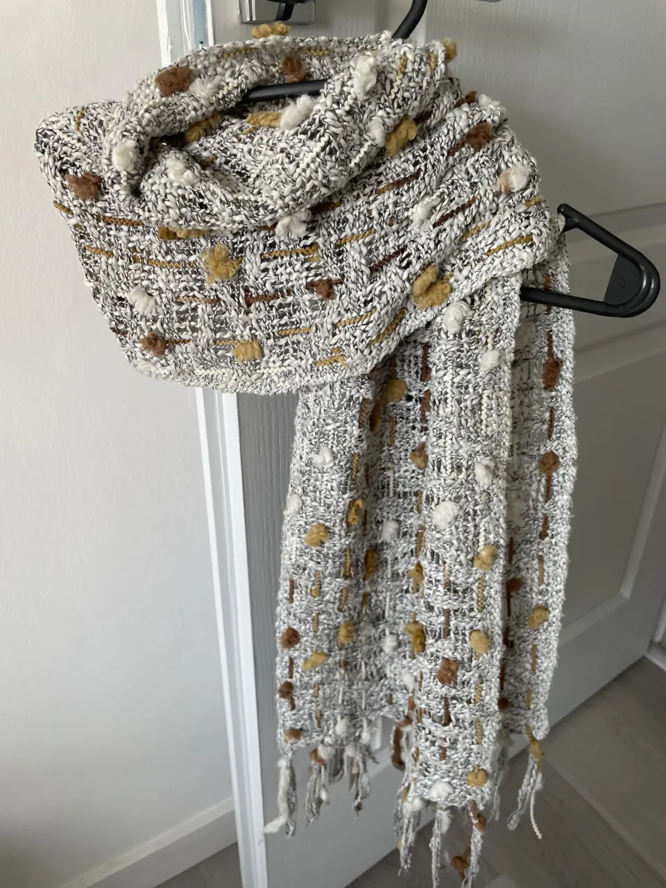 Bufanda tejida con textura irregular y detalles de pompones en tonos crudo, blanco, marrón y dorado. Posee flecos en los extremos.