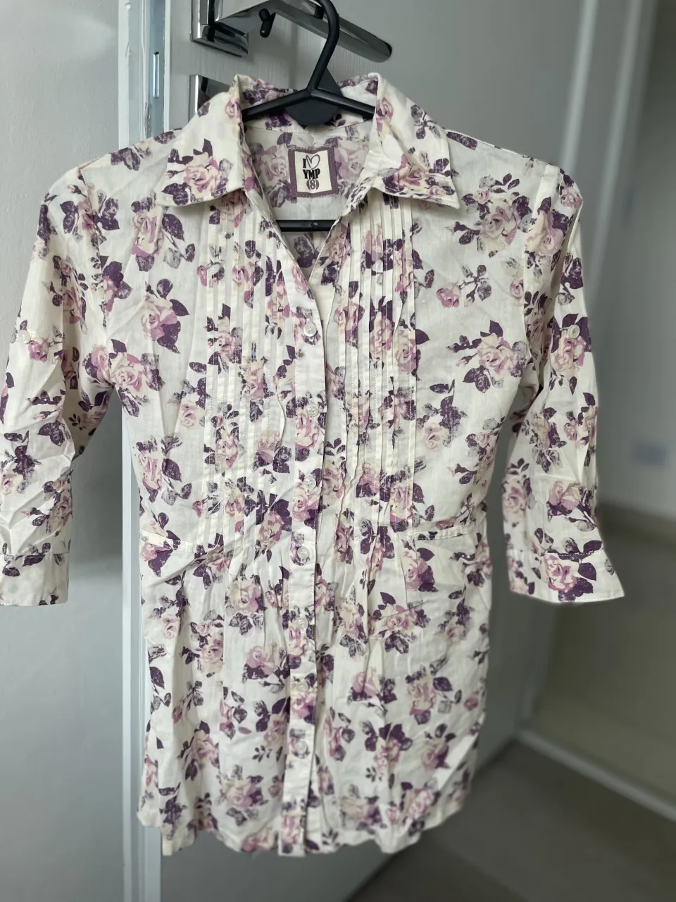 Camisa blanca con estampa floral en tonos lilas y rosas. Posee cuello de camisa, mangas 3/4 y detalles de frunces en el frente. Se abotona al frente y tiene un lazo en la espalda para ajustar.