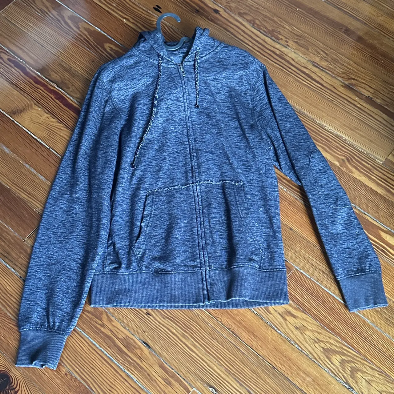 Buzo con cierre y capucha para hombre, de color gris jaspeado. Posee bolsillos delanteros y coderas. Ideal para un look casual y cómodo.