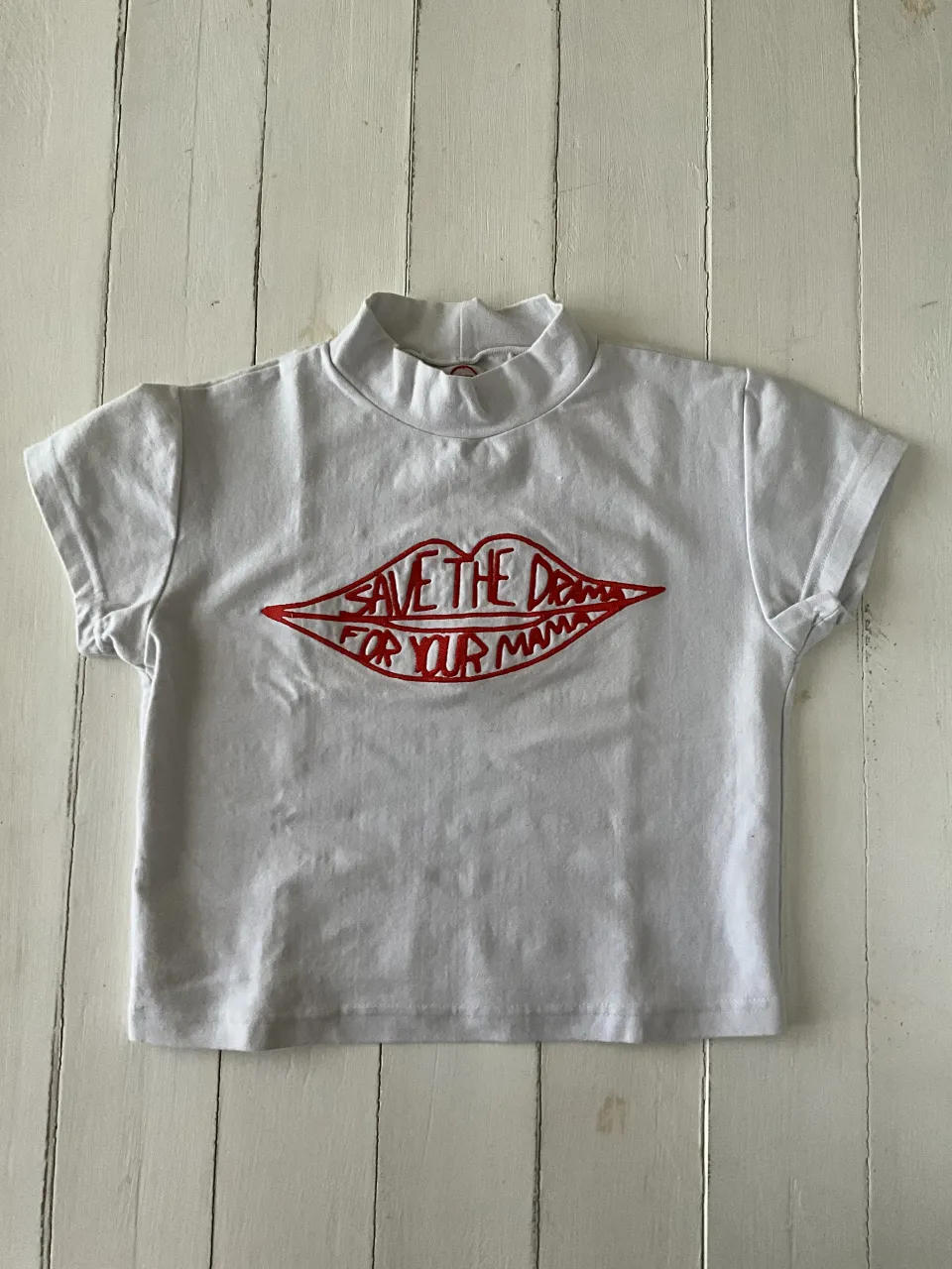 Remera blanca de cuello alto con estampa de labios rojos y la frase "SAVE THE DRAMA FOR YOUR MAMA" en el frente. En la espalda, a la altura del cuello, tiene un pequeño logo bordado en rojo con la palabra "NINJA".