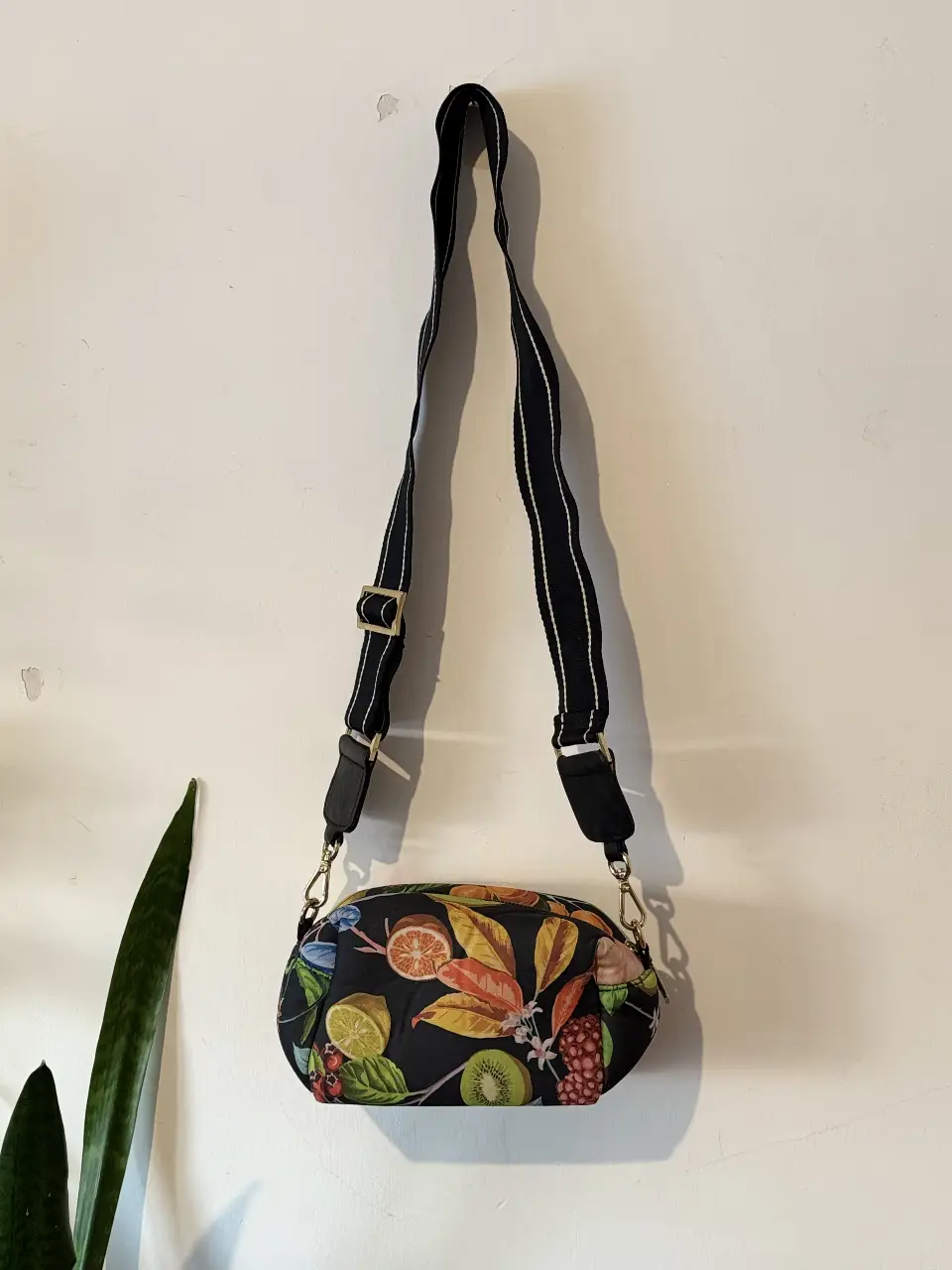 Cartera bandolera con estampado floral y de frutas. Tiene correa ajustable con detalles en blanco y negro. Cuenta con cierre y un interior espacioso.