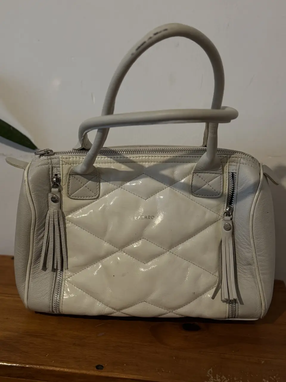 Cartera LAZARO blanca, con diseño de rombos acolchados. Posee dos manijas cortas y una correa larga para llevar al hombro. Detalles de cierres y borlas en los laterales. Ideal para un look elegante y sofisticado.