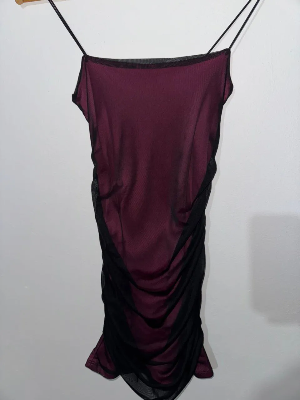 Vestido corto de tirantes finos, con tela de red negra superpuesta sobre una base de color bordó. Presenta fruncido lateral.