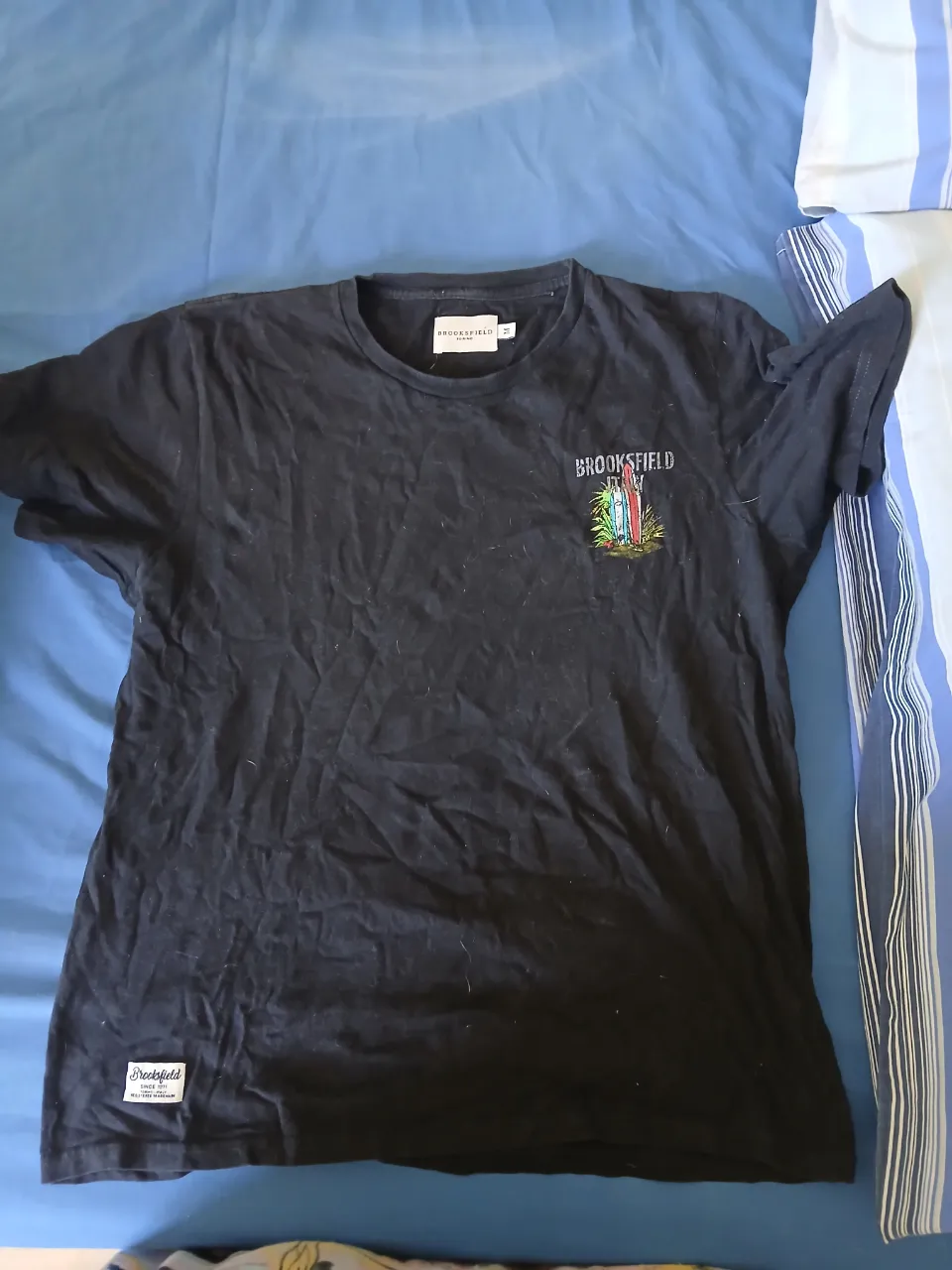 Remera negra de manga corta con estampa de tablas de surf y palmeras en el frente. Posee un logo de la marca en la parte inferior izquierda.