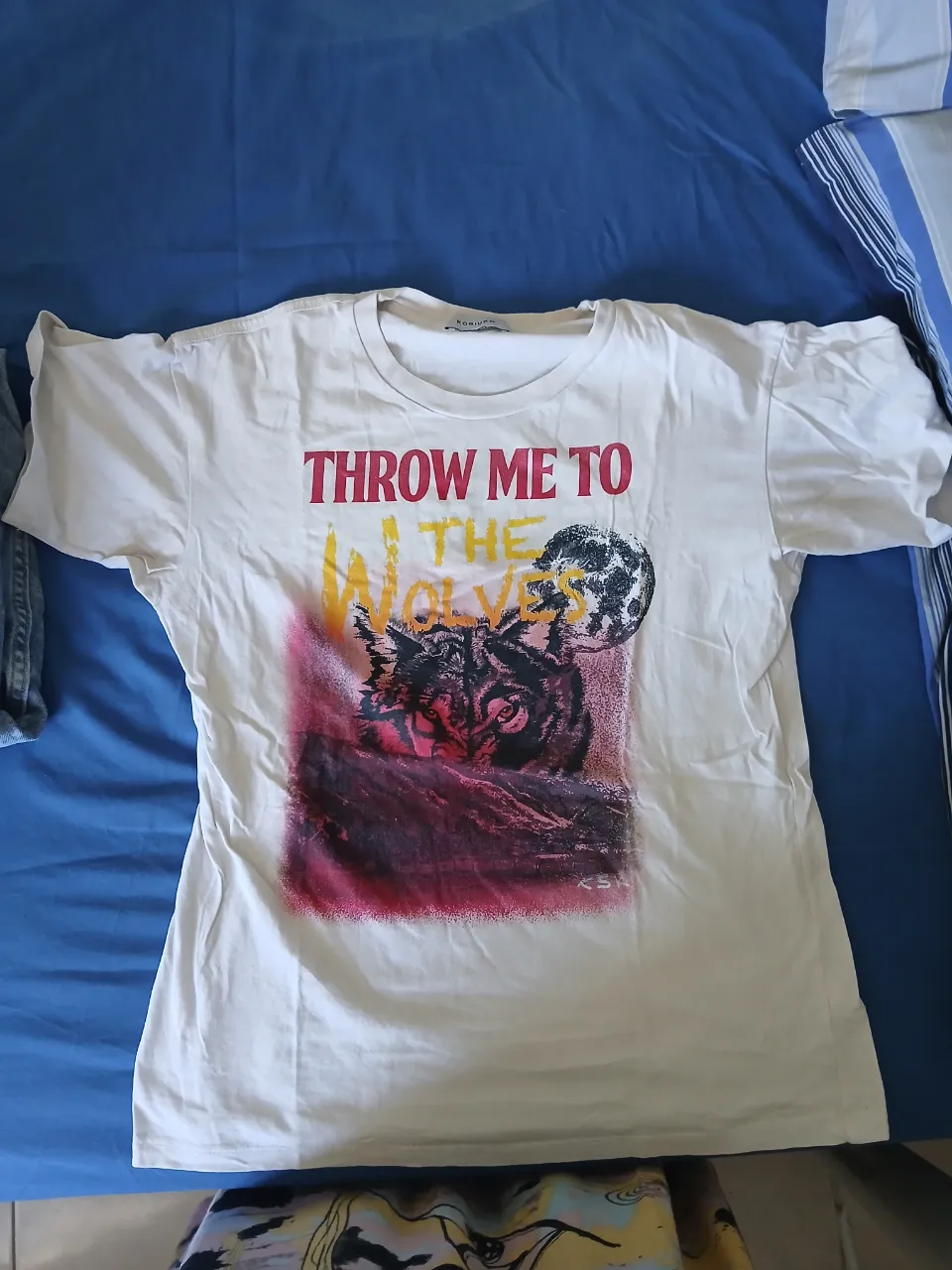 Remera blanca con estampa de lobo y luna. La estampa tiene colores rojizos, amarillos y negros. En la parte superior de la estampa se lee "THROW ME TO THE WOLVES".
