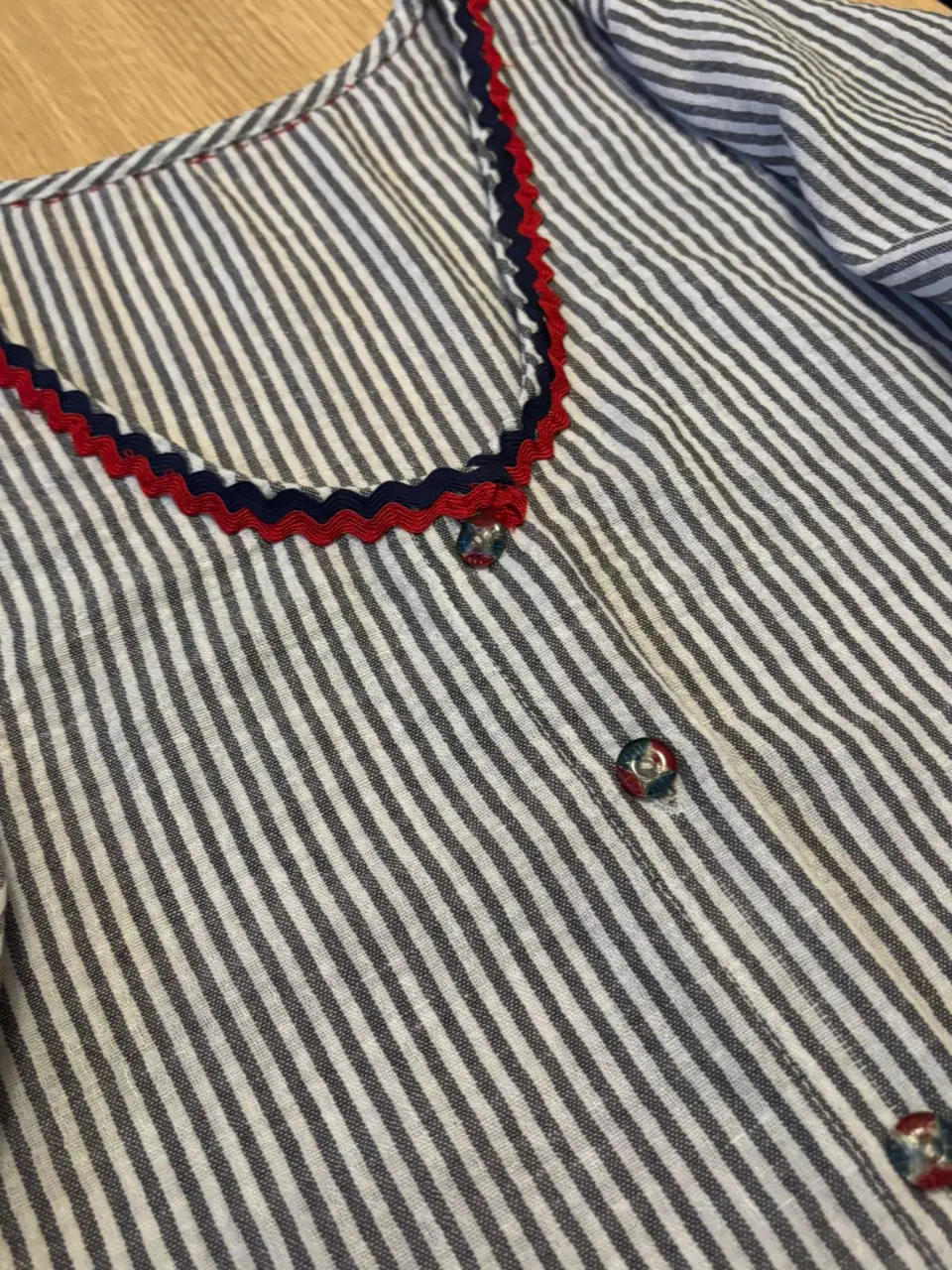 Blusa a rayas verticales en blanco y azul, con detalles en rojo y azul marino en el cuello y botones. Mangas 3/4 con puños y botones. Ideal para un look casual y fresco.