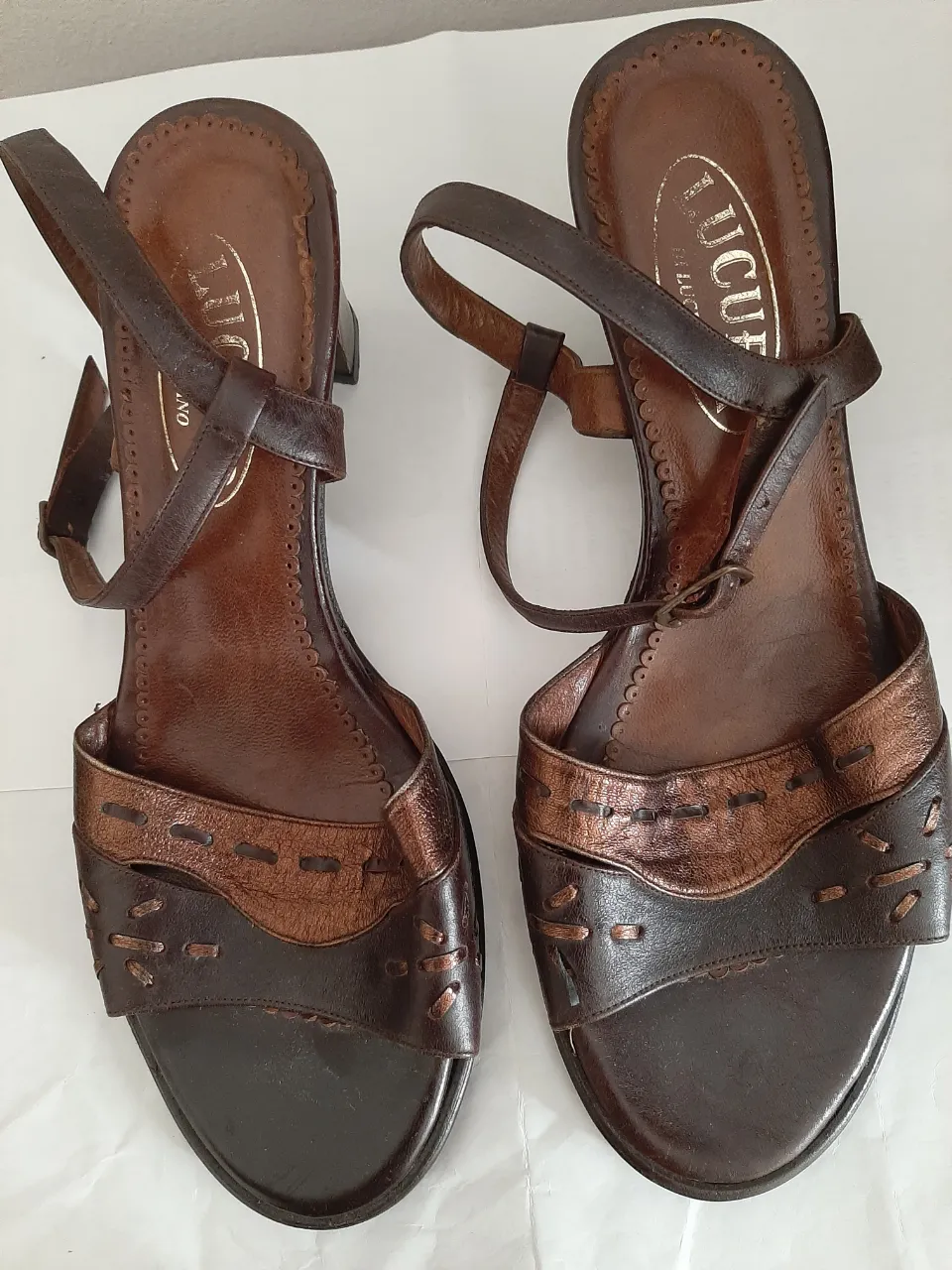 Sandalias de cuero marrón con detalles metálicos y costuras a la vista. Tienen tiras finas que se cruzan en el empeine y una tira con hebilla en el tobillo. Taco grueso y cómodo.