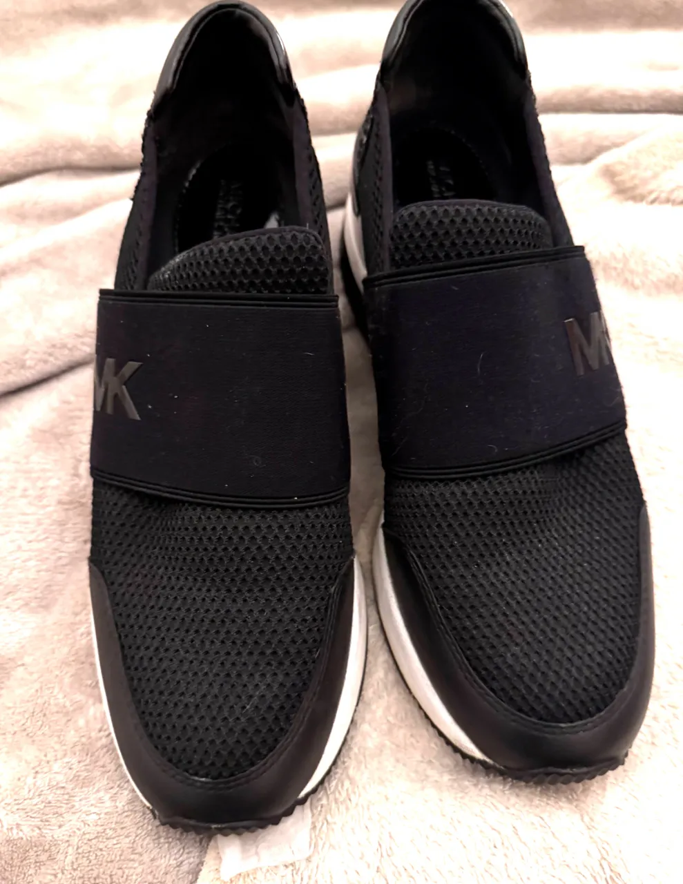 Zapatillas deportivas negras de mujer, con detalles en blanco y logo MK en la banda elástica. Ideales para un look casual y cómodo.