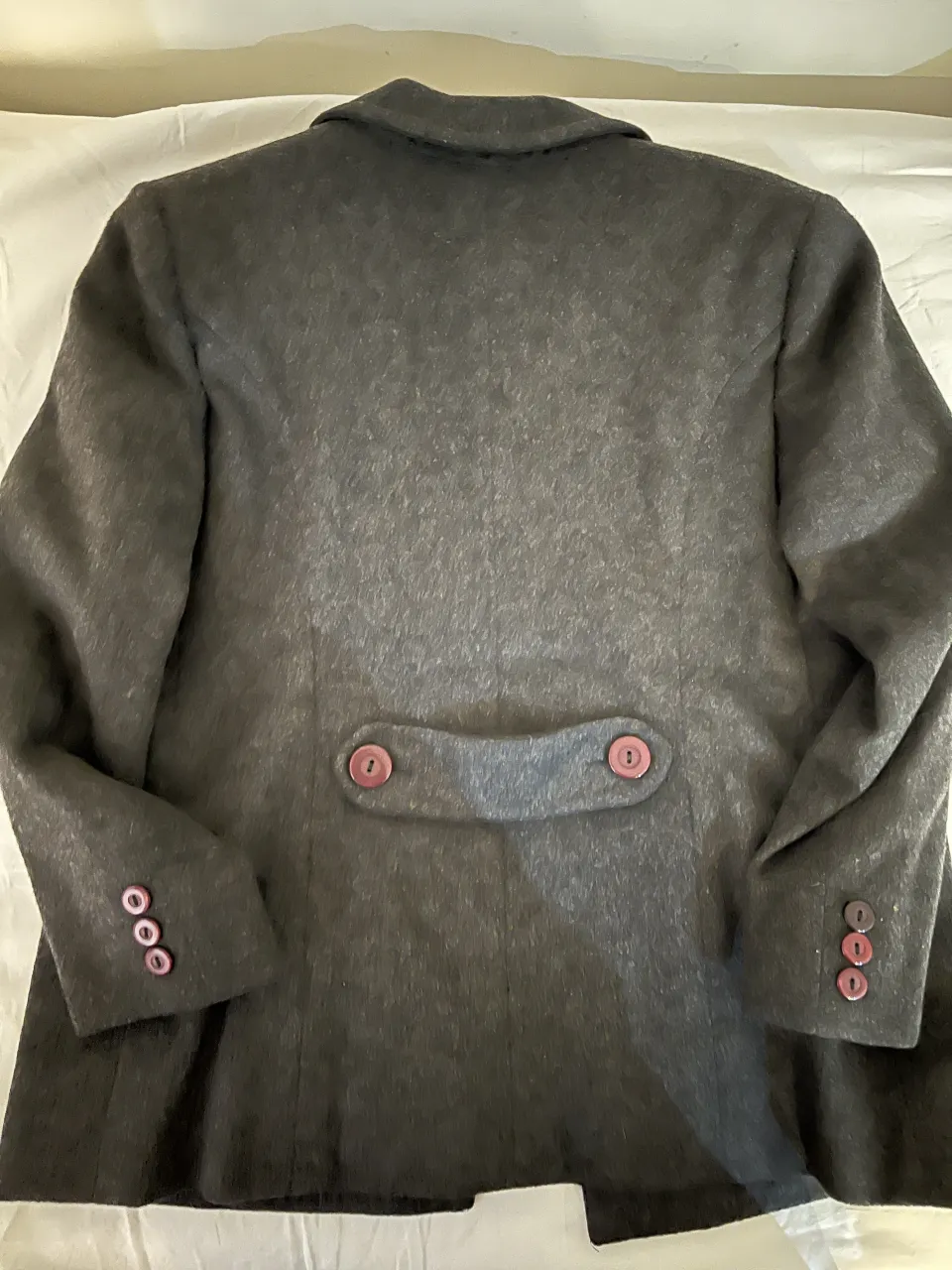 Abrigo de paño gris oscuro con botones fucsia. Tiene dos filas de botones en el frente y tres botones en cada puño. En la parte trasera, presenta un detalle de solapa con dos botones.