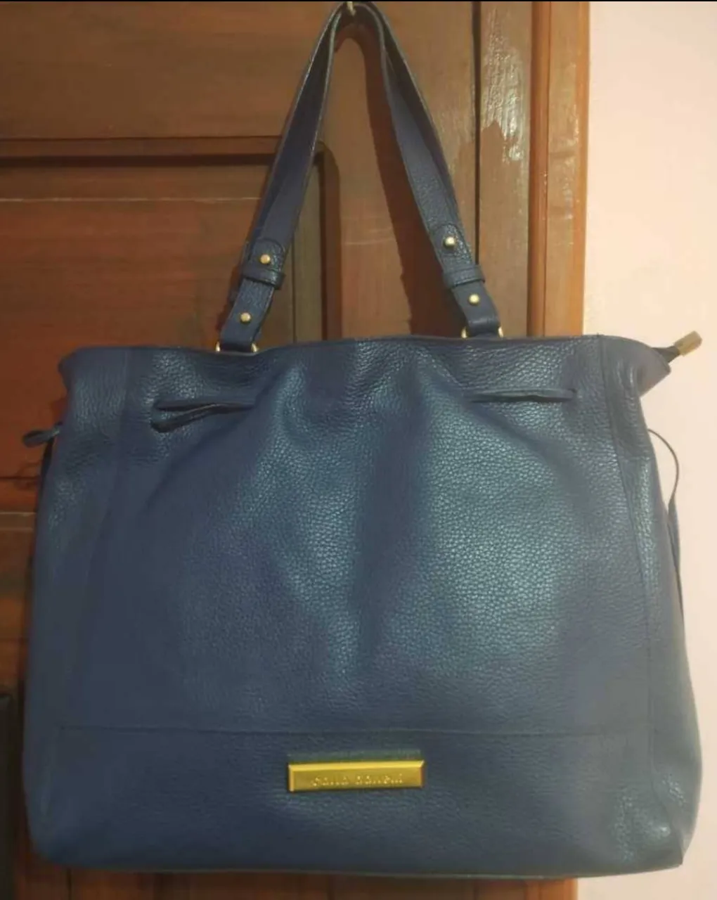 Bolso grande de cuero azul marino con cierre y un cordón ajustable en la parte superior. El interior es de tela y tiene tres bolsillos (uno con cierre). Viene la bolsa antipolvo original. Las medidas son: ancho arriba- 39, ancho en la base- 35. Altura: 31 cm y profundidad: 10 cm. Sin imperfecciones.