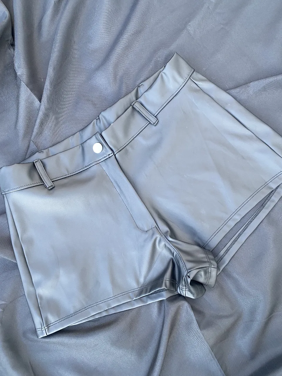 Short de cuero sintético negro, tiro bajo, con detalles de costura y cierre con botón. Ideal para un look moderno y audaz.