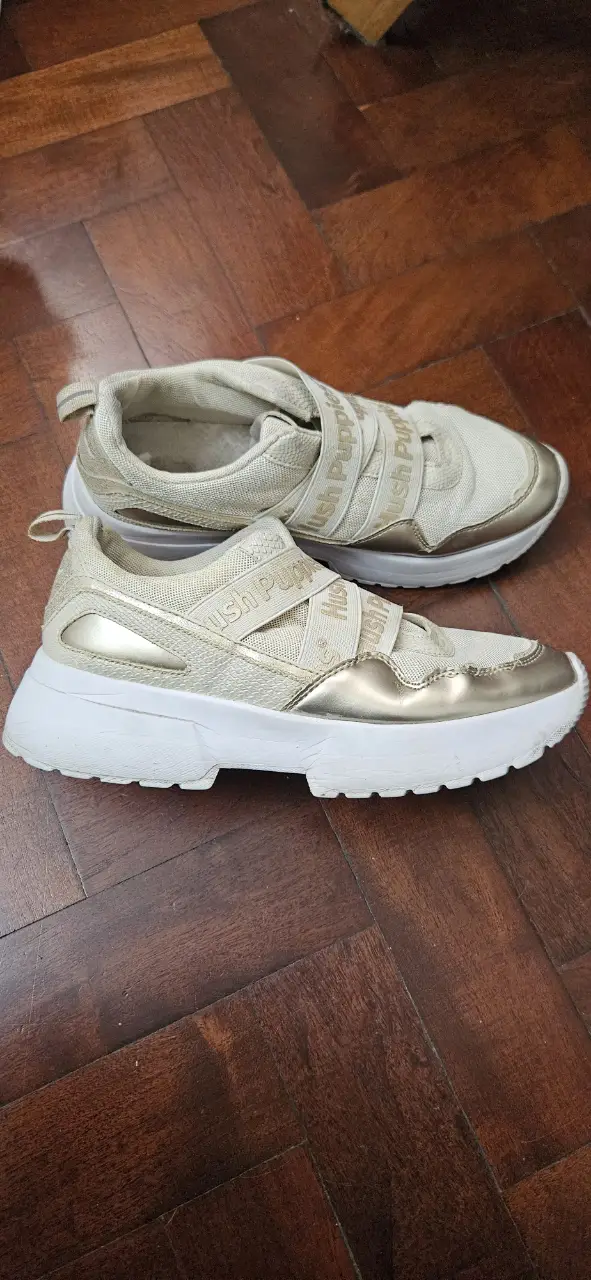 Zapatillas Hush Puppies doradas con detalles blancos. Tienen tiras con el logo de la marca y suela gruesa. Ideales para un look casual y cómodo.
