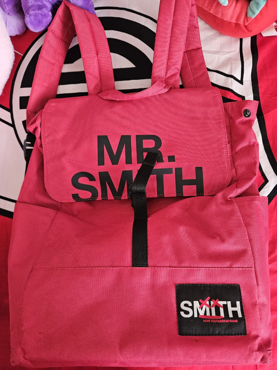 Mochila rojo vibrante con estampa "MR. SMITH" en negro en el frente. Tiene un bolsillo frontal con cierre y un logo de "SMITH" en la parte inferior. Ideal para el día a día o para llevar tus esenciales a todos lados con estilo.
