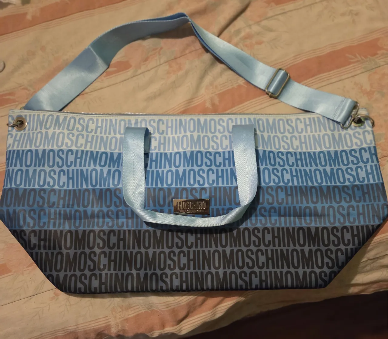 Bolso tote de mujer Moschino con estampado repetido del logo en tonos azules. Cuenta con manijas cortas y correa larga ajustable para llevar al hombro. Ideal para llevar tus pertenencias con estilo y comodidad.