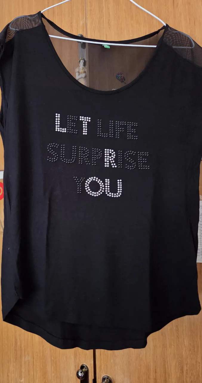 Remera negra de mujer con frase "LET LIFE SURPRISE YOU" estampada con brillos. Cuello redondo y mangas cortas con detalle de tul en los hombros. Ideal para un look casual y moderno.