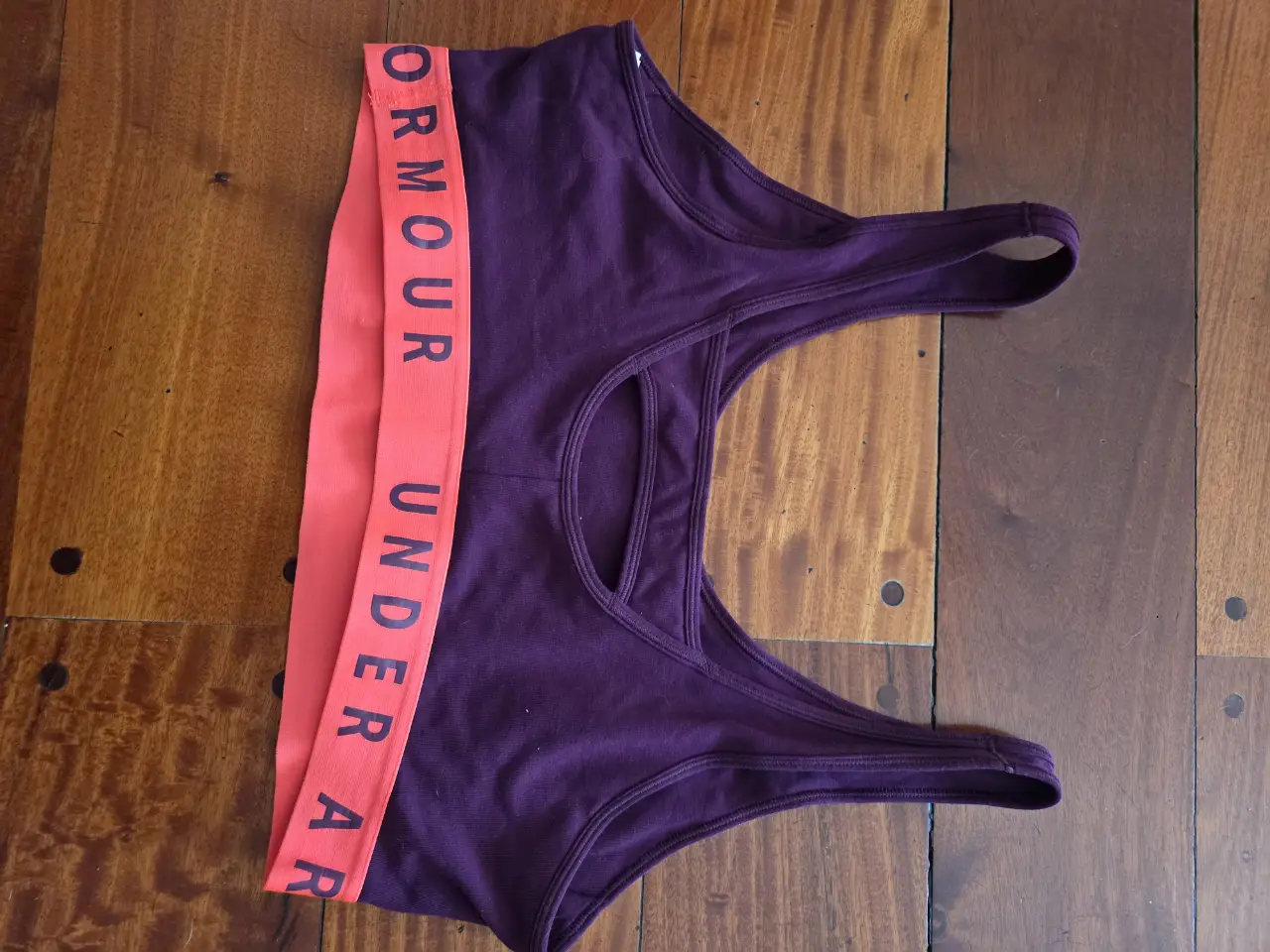 Top deportivo Under Armour en color violeta con banda en color naranja con el logo de la marca. Ideal para entrenamientos de alto impacto.