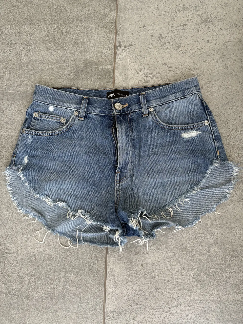 Shorts de jean tiro alto con roturas y detalles deshilachados en el borde inferior. Cierre con botón y cierre.