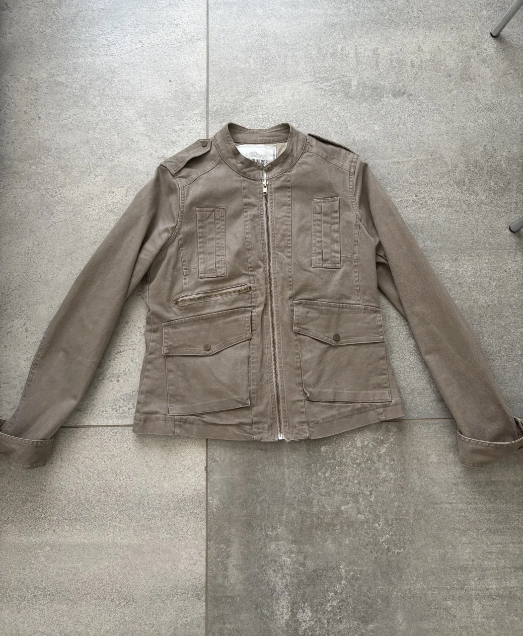 Campera de tela color tiza con cuello mao, cierre completo al frente y bolsillos con tapa en el frente. Tiene hombreras y puños con botón.