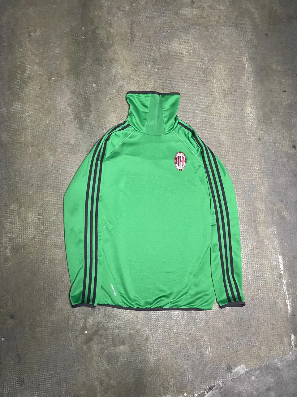 Buzo deportivo de cuello alto del AC Milan, con las tres tiras negras de Adidas en las mangas. Cuenta con el logo del equipo y la marca Adidas.