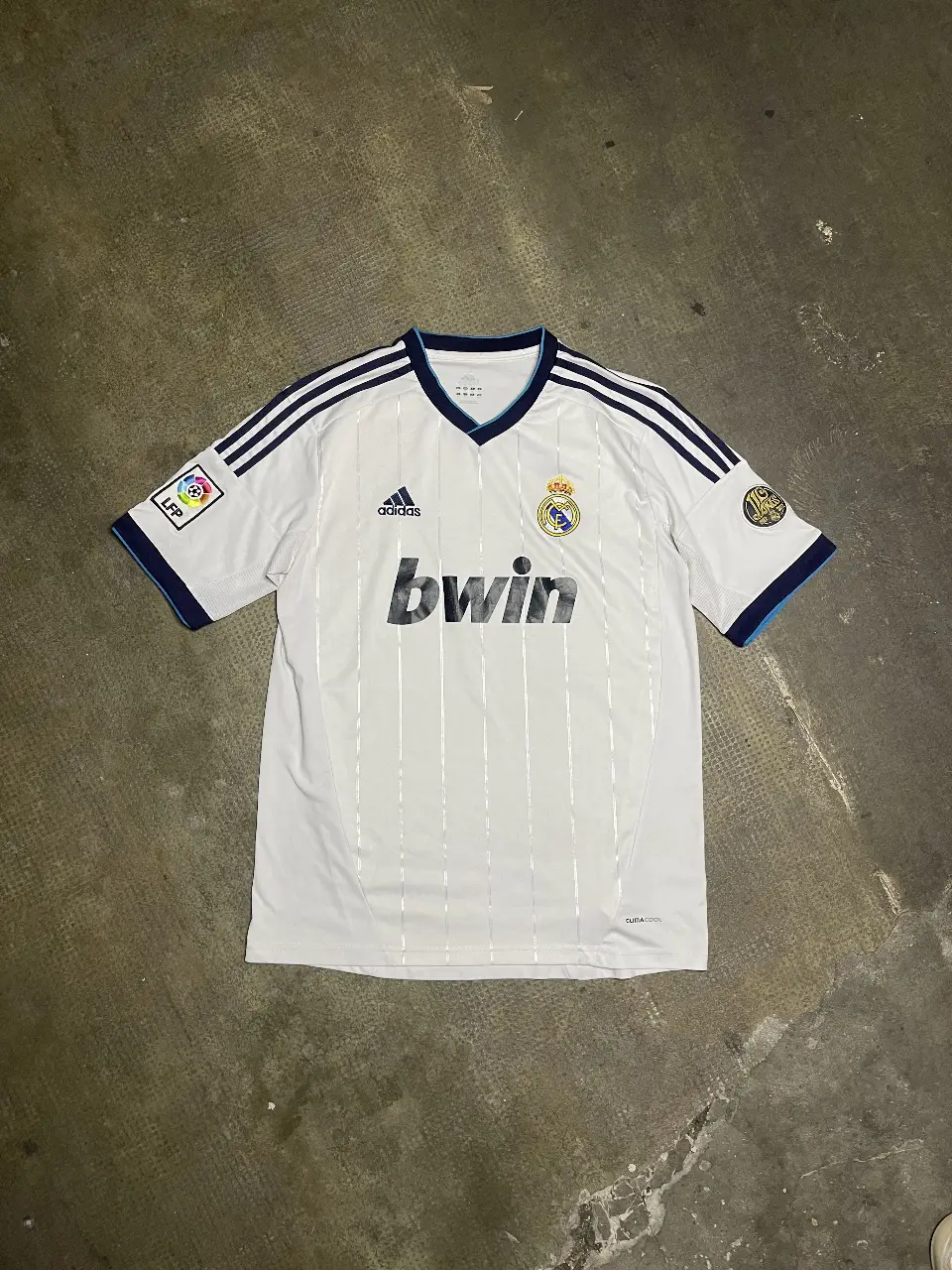 Camiseta de fútbol del Real Madrid, blanca con detalles en azul y el logo de Adidas y bwin. Cuenta con el escudo del club y parches en las mangas. Ideal para fanáticos y coleccionistas.