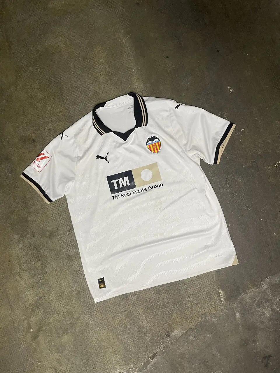 Camiseta de fútbol del Valencia CF, blanca con detalles en negro y dorado. Con el logo de Puma y el sponsor TM Real Estate Group. Ideal para fanáticos y coleccionistas.