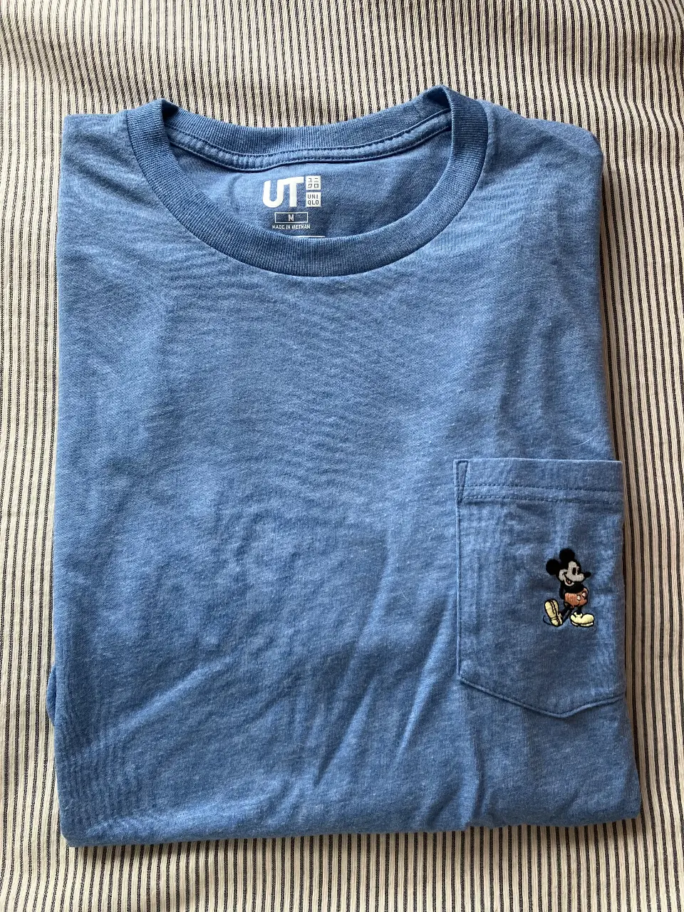 Remera de manga corta, color azul claro, con un bolsillo en el pecho con un bordado de Mickey Mouse. La remera es de la marca Uniqlo.