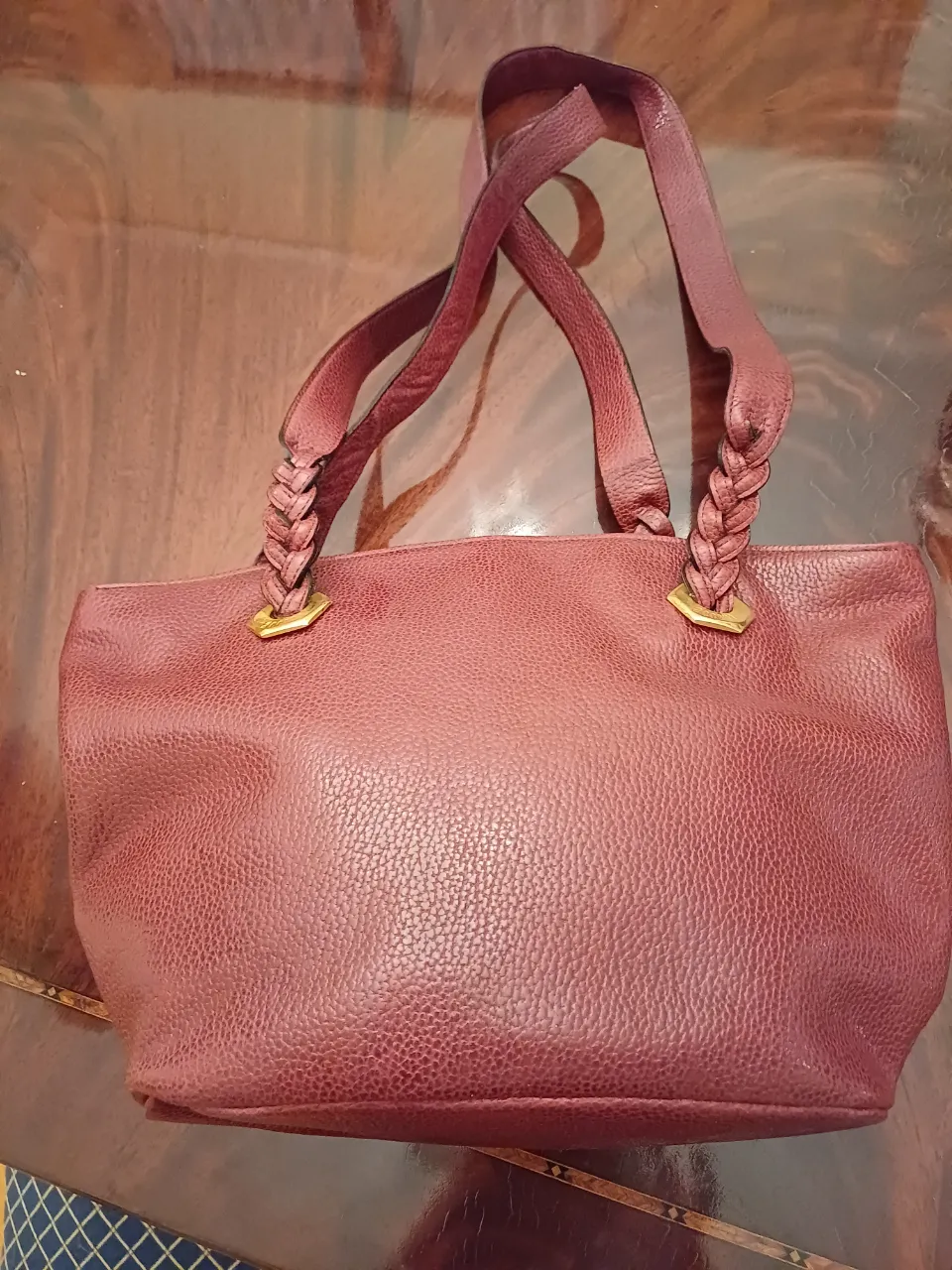 Cartera de mano de cuero color borgoña con detalles trenzados en las asas y herrajes dorados. Ideal para un look elegante y sofisticado.