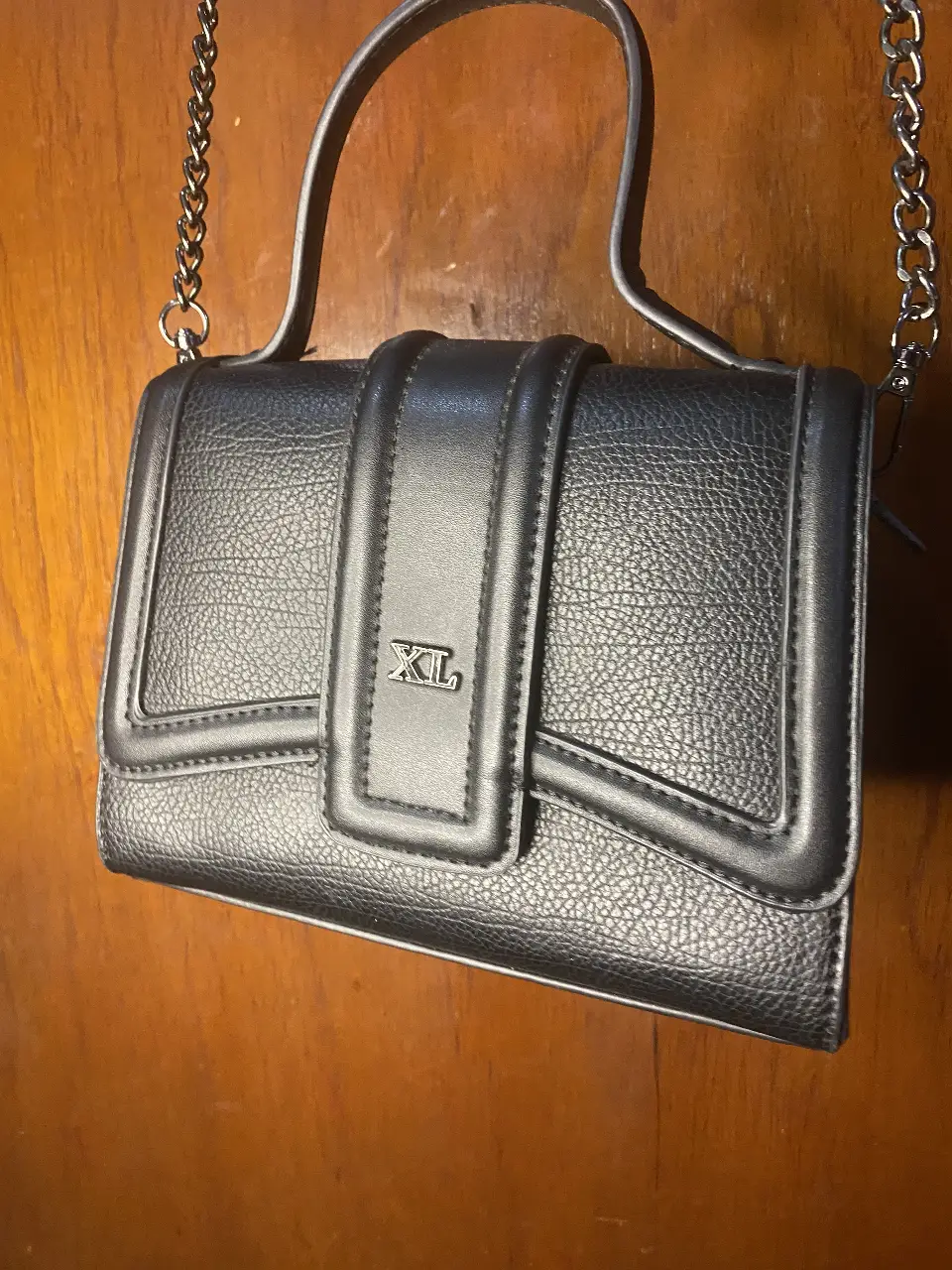 Cartera negra con correa de cadena y manija superior. Diseño elegante con detalle XL en el frente. Ideal para salidas y eventos.
