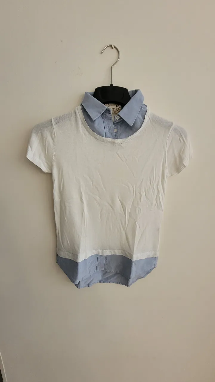 Remera blanca de manga corta con cuello de camisa celeste. La parte inferior de la prenda es celeste y simula ser una camisa.